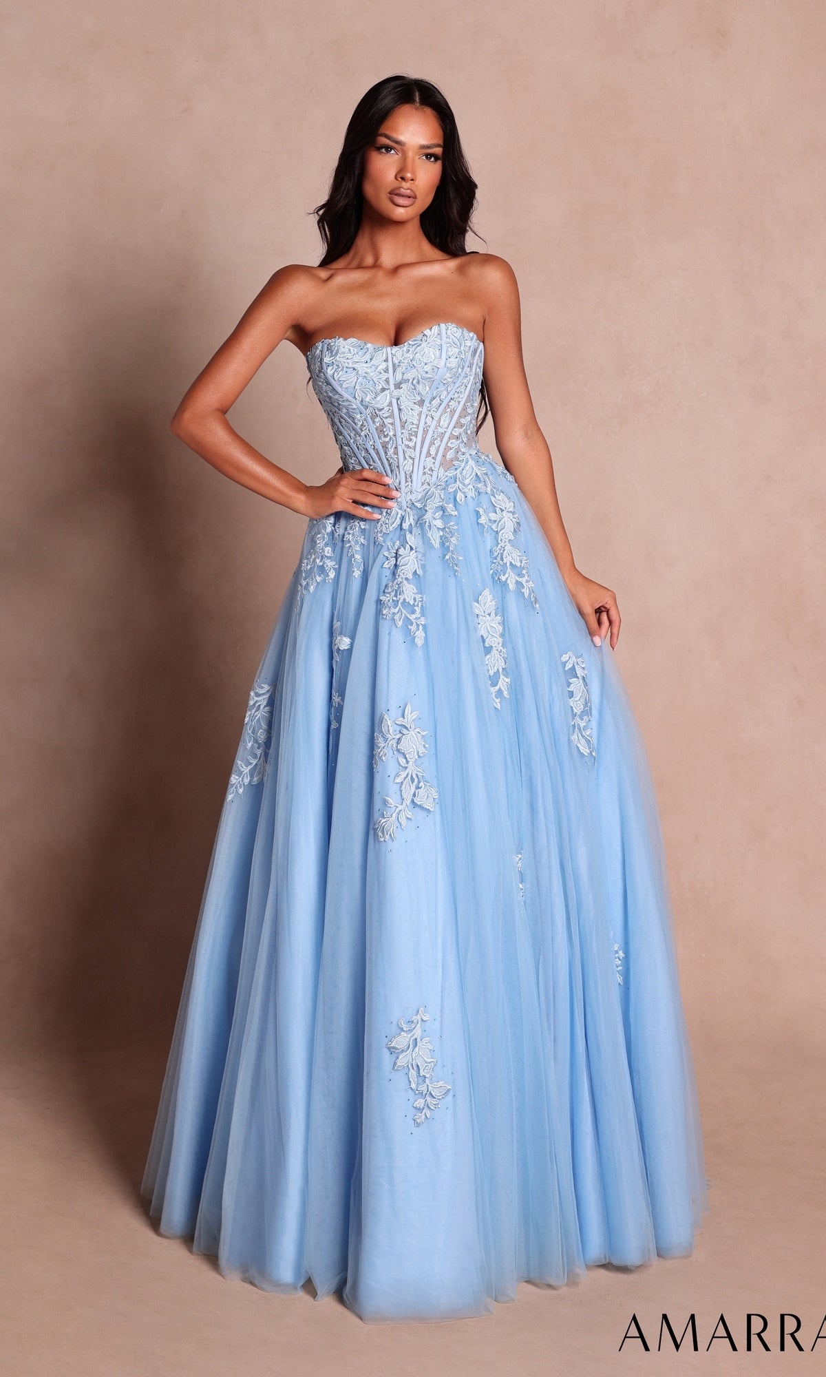 A woman stands against a plain beige background wearing the Amarra 89626 long prom dress in light blue—a strapless ball gown featuring floral lace appliqué and a fitted bodice with a sweetheart neckline.